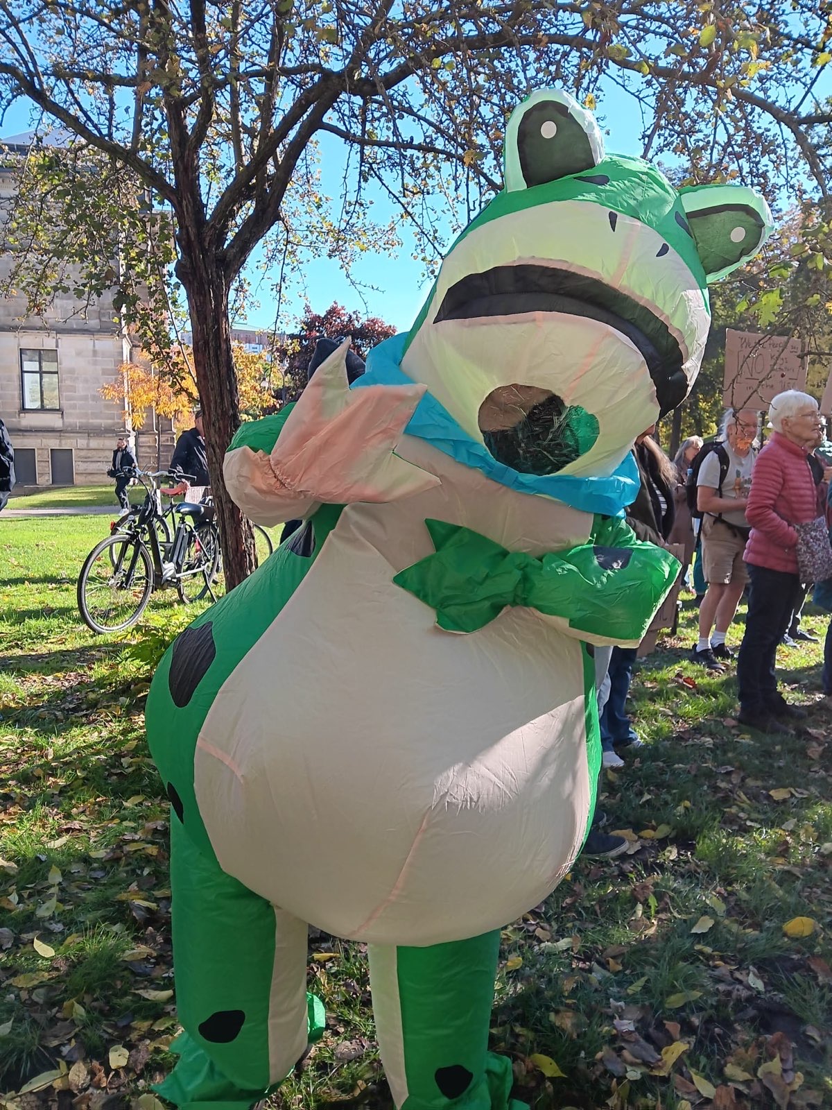 A big green inflatable frog is waving into the camera. A group of protesters is standing in a sunny park in the background.
