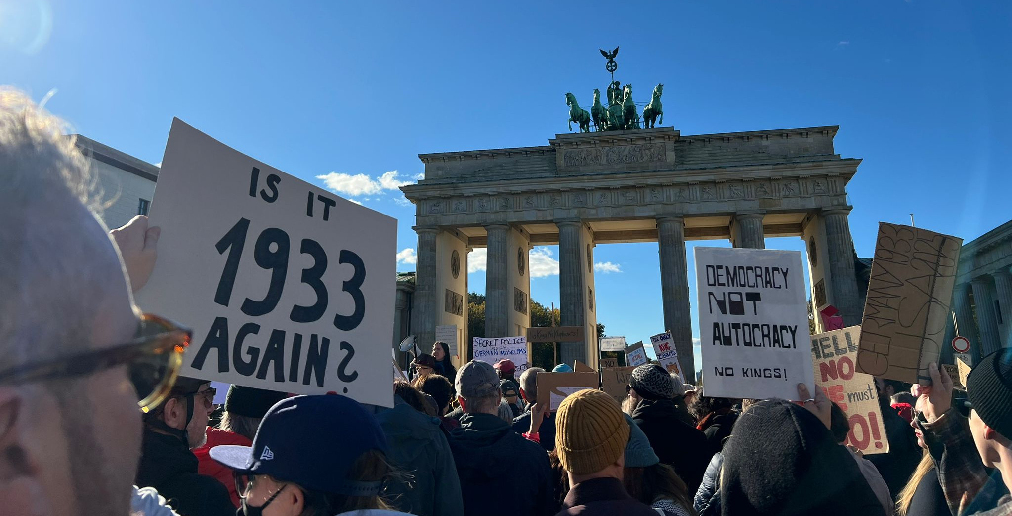 A group of people protesting in front of the Brandenburg gate. Some of them are holding signs. One sign in the front reads 'Is it 1933 again'. Another sign says 'Democracy Not Autocracy - No Kings'.